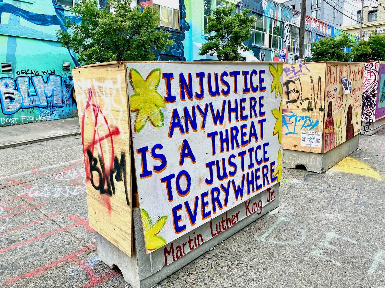 A row of concrete traffic barricades are covered in plywood and painted with a variety of messages. This is in the Black Lives Matter protest area of Capitol Hill in Seattle. This sign says, Injustice anywhere is a threat to justice everywhere, a quote by Martin Luther King Jr.