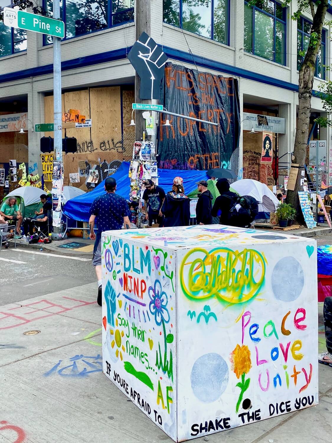 Signage at the CHOP area of Seattle during the Black Lives Matter protest. This is a square box created from plywood sitting in front of the vacant East Precinct police station. The colors of the box are white background with hints of light green and blue. The sign says, Peace, Love, Unity.