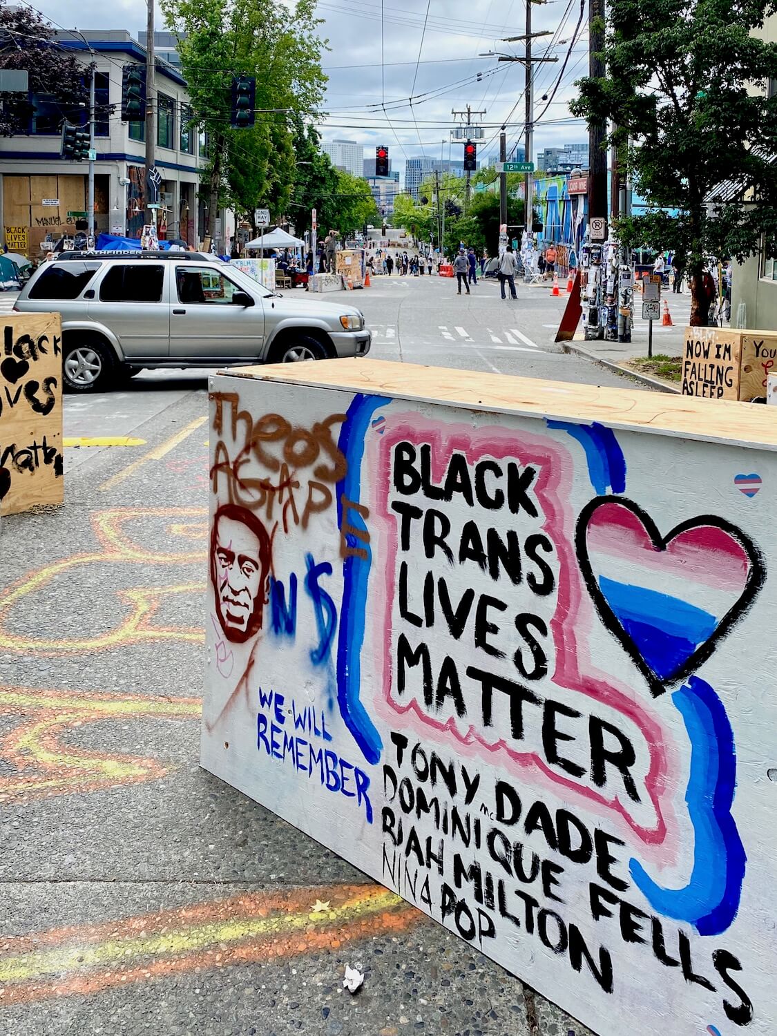 Signage at the CHOP area of Seattle during the Black Lives Matter protest. The colors are a bright mix of blues and greens and this sign says, Black Trans Lives Matter, with the list of four names of those trans people killed by police.