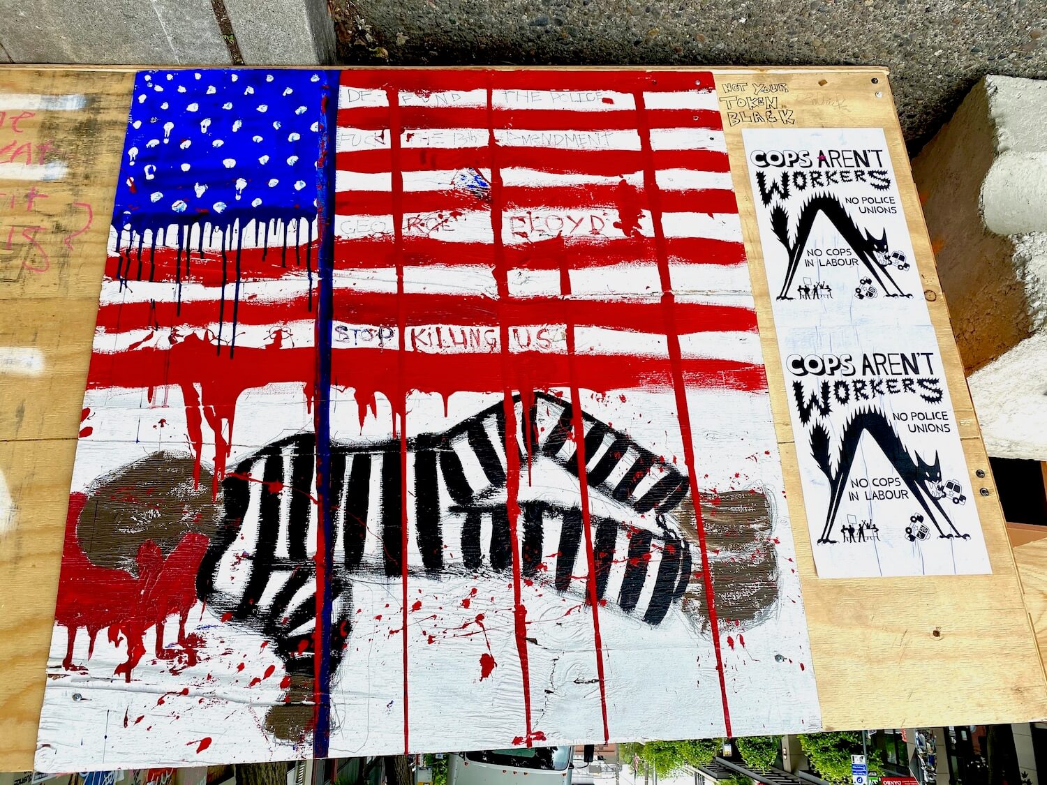 Signage at the CHOP area of Seattle during the Black Lives Matter protest. Red white and blue paint makes out a dripping American flag with a dead man in striped prison outfit lies below, blood gushing from his head. The words say, Stop Killing US.