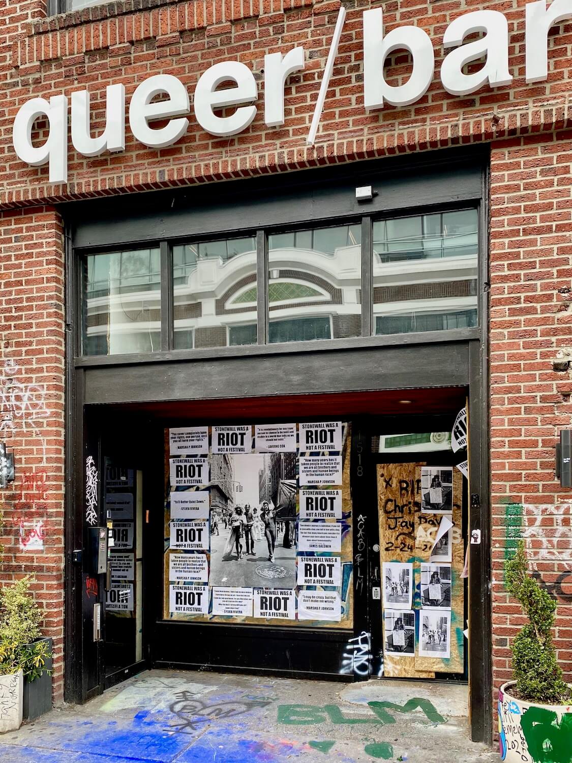 The front of Queer Bar, in the are of Black Lives Matter protest zone in Seattle.  The front of the bar is red brick and the doorway is matted with white posters that say, Stonewall was a Riot, not a festival.  