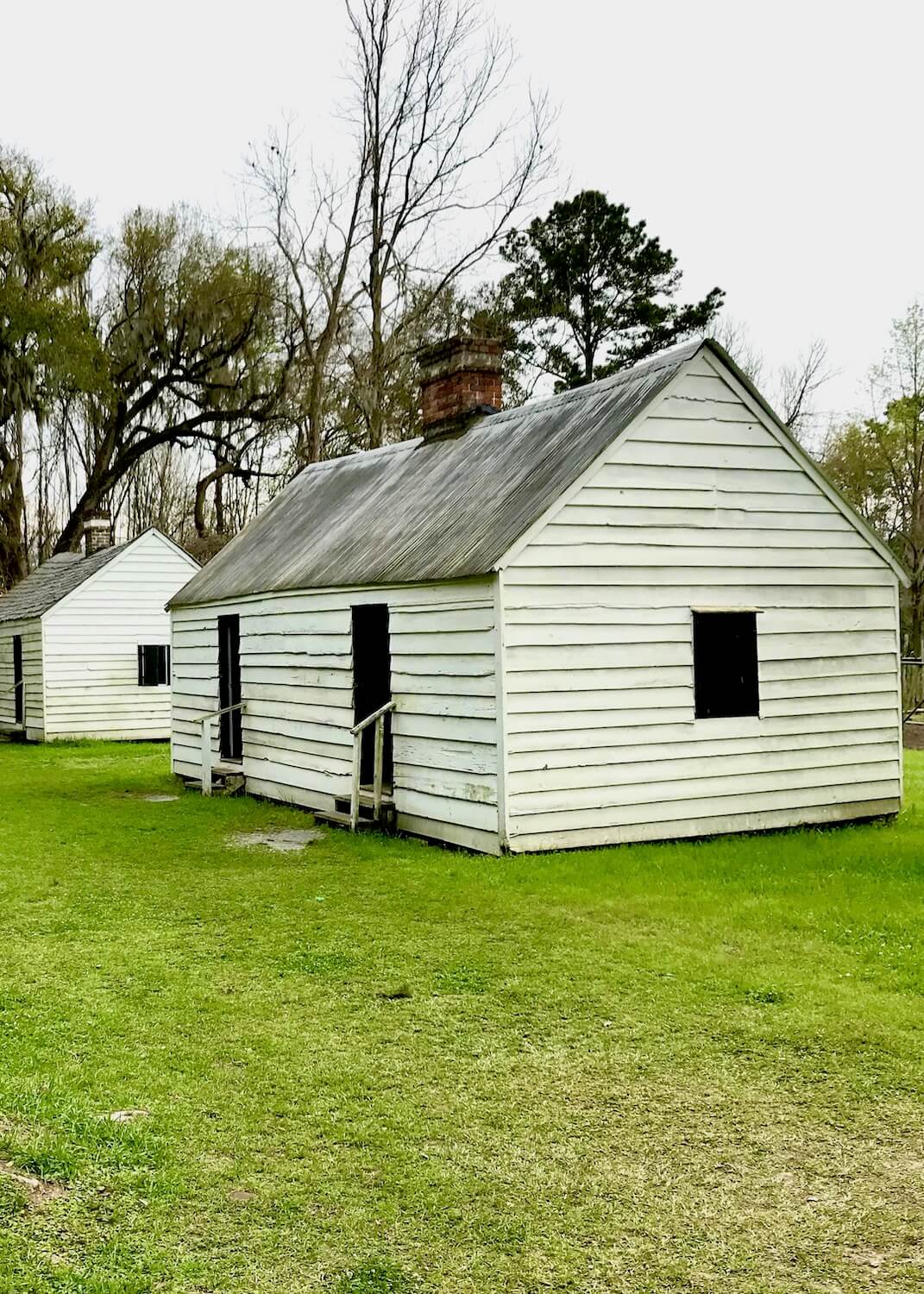 Two simple cabins that stand to represent the housing conditions of slaves on a rice plantation near Charleston, SC.  The rectangular buildings are made of white painted narrow wood planks and variegated gray sheet metal with a simple red brick chimney in the middle.  There are a few dark openings for window areas and two doorways in the front with steps leading to the green grassy area outside.  