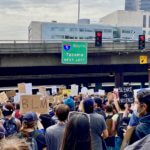 A large crowd marches down a steep roadway in Seattle, Washington towards a bridge with an interstate sign directing traffic South to Tacoma and two red stoplights. The people are holding various signs that promote Black Lives Matter and other slogans like Silence is Violence. In the background are other city buildings, including a sliver of the tan colored municipal jail.
