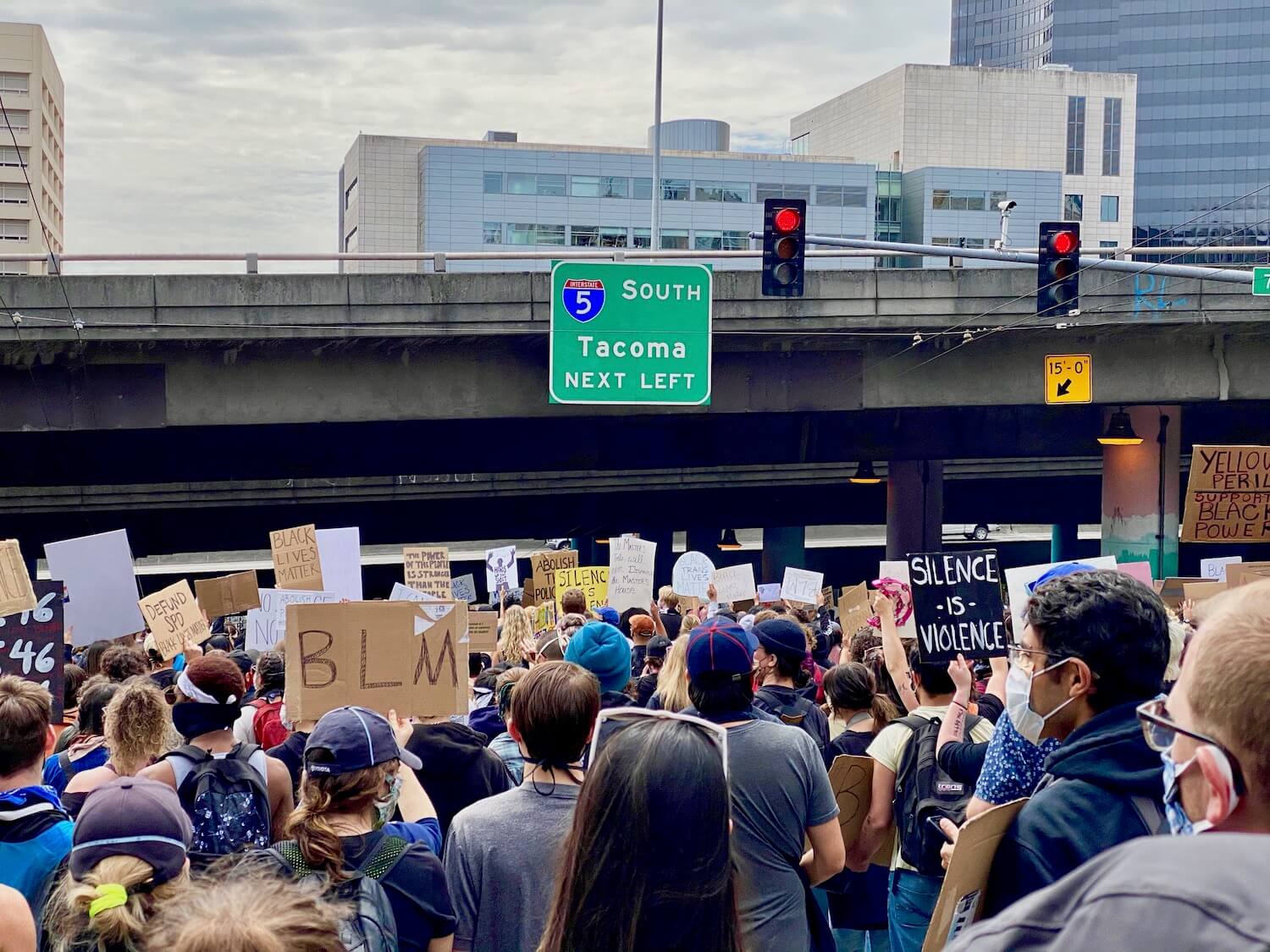 A large crowd marches down a steep roadway in Seattle, Washington towards a bridge with an interstate sign directing traffic South to Tacoma and two red stoplights.  The people are holding various signs that promote Black Lives Matter and other slogans like Silence is Violence.  In the background are other city buildings, including a sliver of the tan colored municipal jail.  