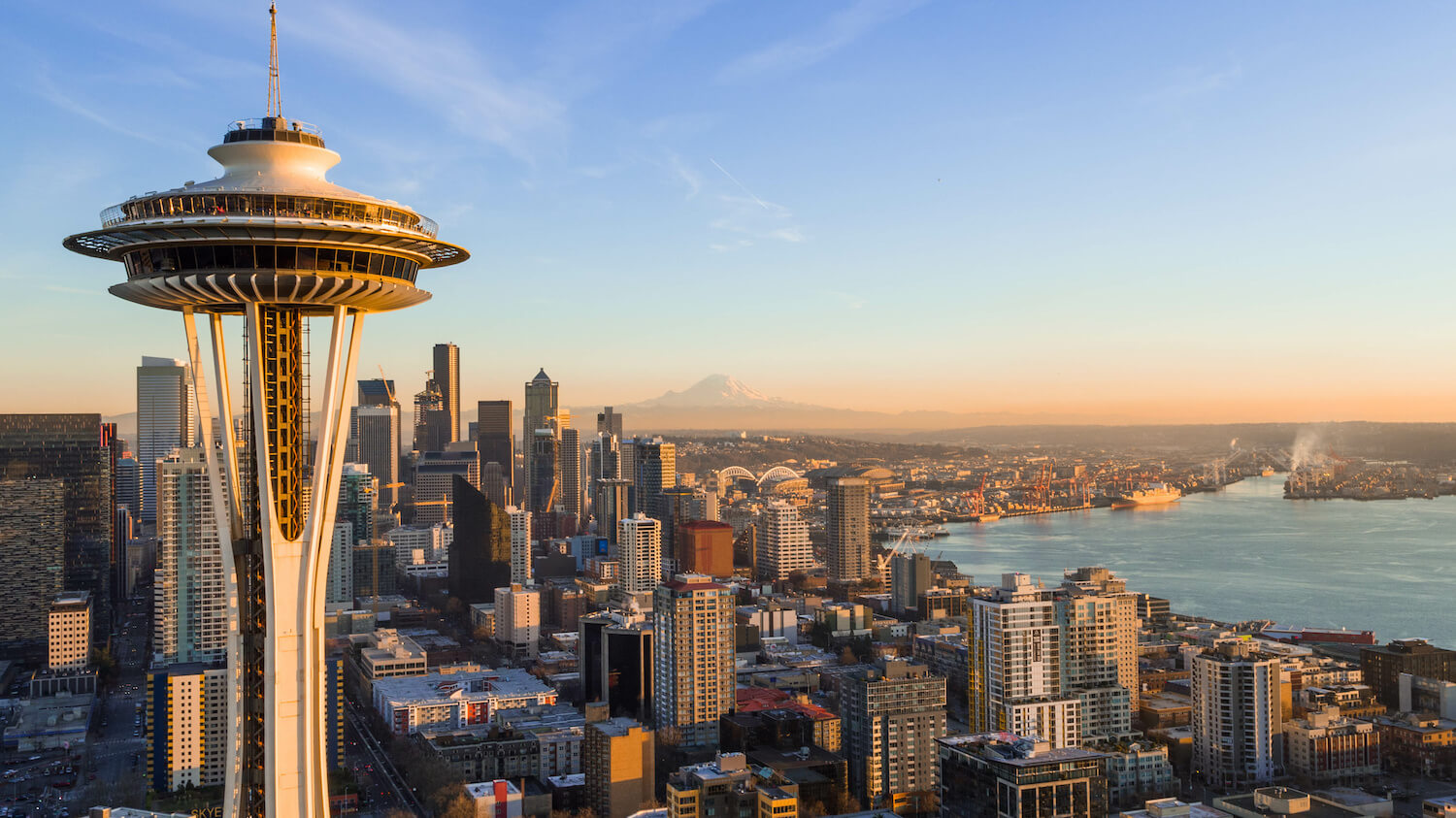 Skyline of Seattle Washington from the point of view of the Space Needle, which is in the foreground of this shot. The many buildings of the Seattle skyline crowd around Elliott Bay with the faint outline of Mt. Rainier on the peach colored horizon.