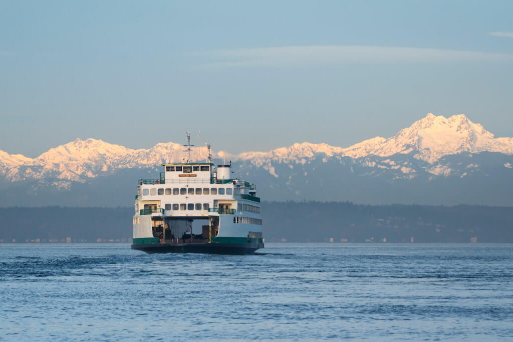 A Washington State Ferry crosses a channel of the Puget Sound, en route to the Olympic Peninsula. The snow capped mountains shine in front of a blue sky and layers of evergreen trees are seen closer to the ferry, which glides through lightly rolling water.
