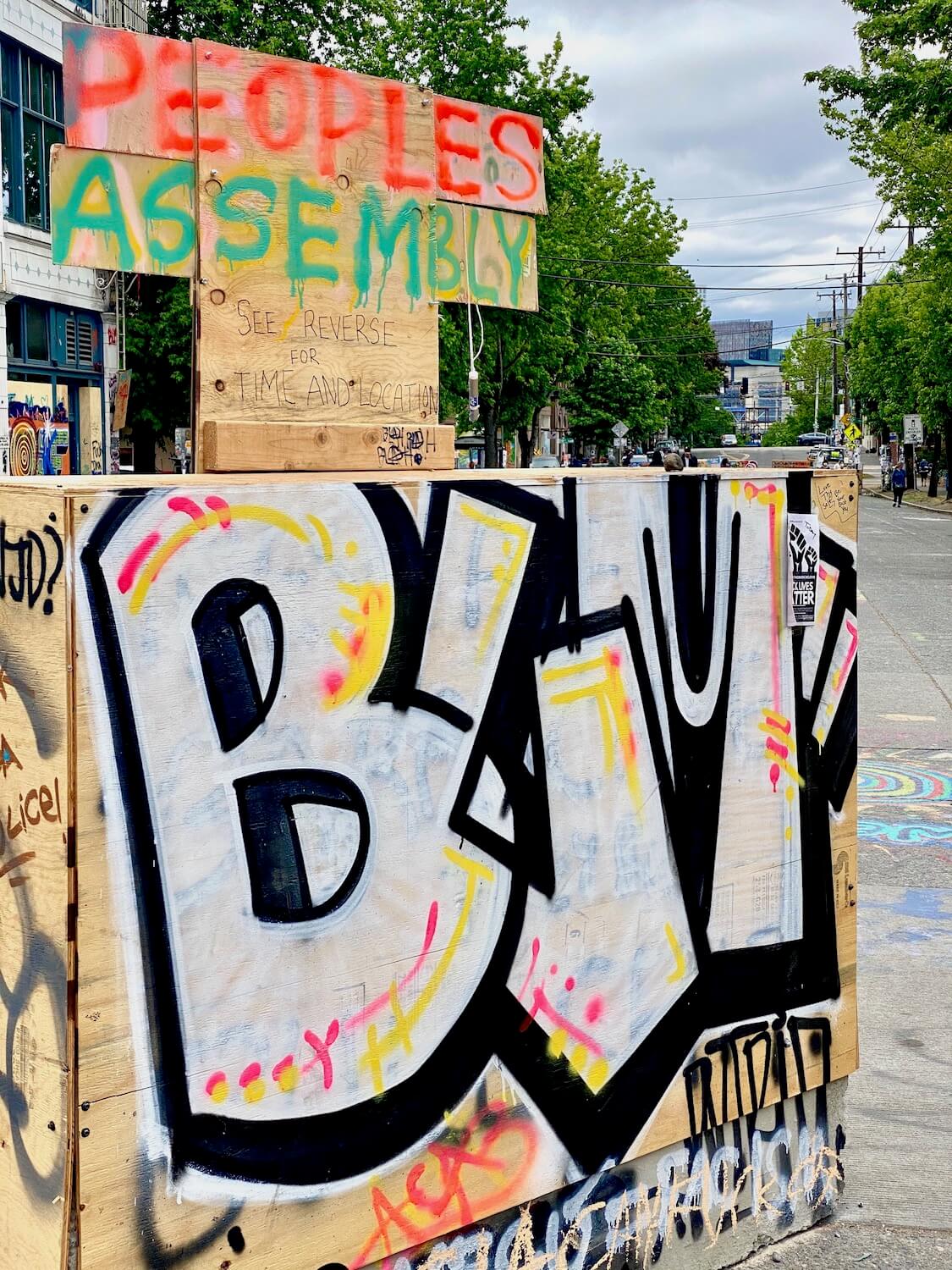 A parking barrier covered in painted plywood shows graffiti letters BLM for Black Lives Matter.  This is one of the  blocks in the protest zone known as CHOP in Seattle. 