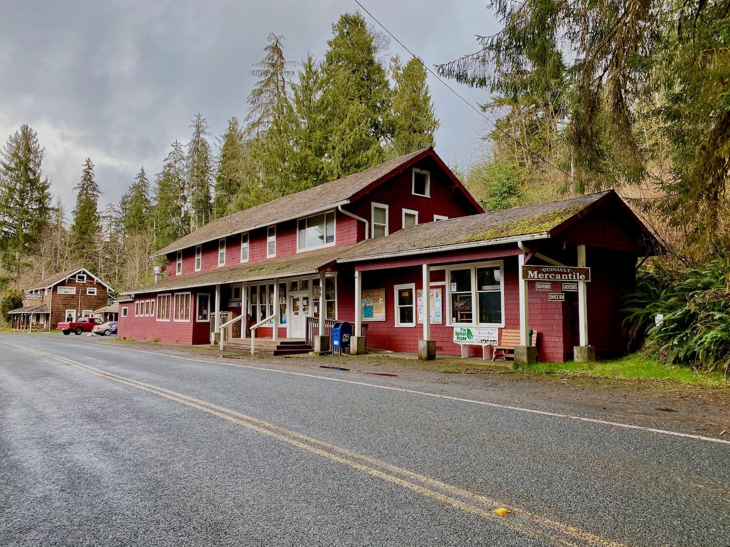 The old mercantile wears bright red paint well and the color pops against white trim. This 1930s era building hugs the side of the main paved road, with a yellow line down the middle. The background shows a gray sky heavy with potential rain.