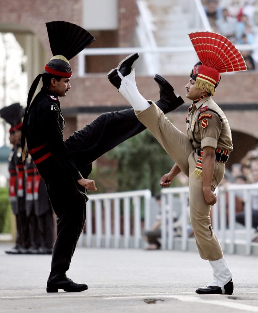 Border guards at Wagah, the checkpoint between India and Pakistan, kick up their legs at one another. The Pakistani guard is wearing black with red accents and a top notch while the Indian guard wears a khaki colored outfit with white boot covers and a red top notch.