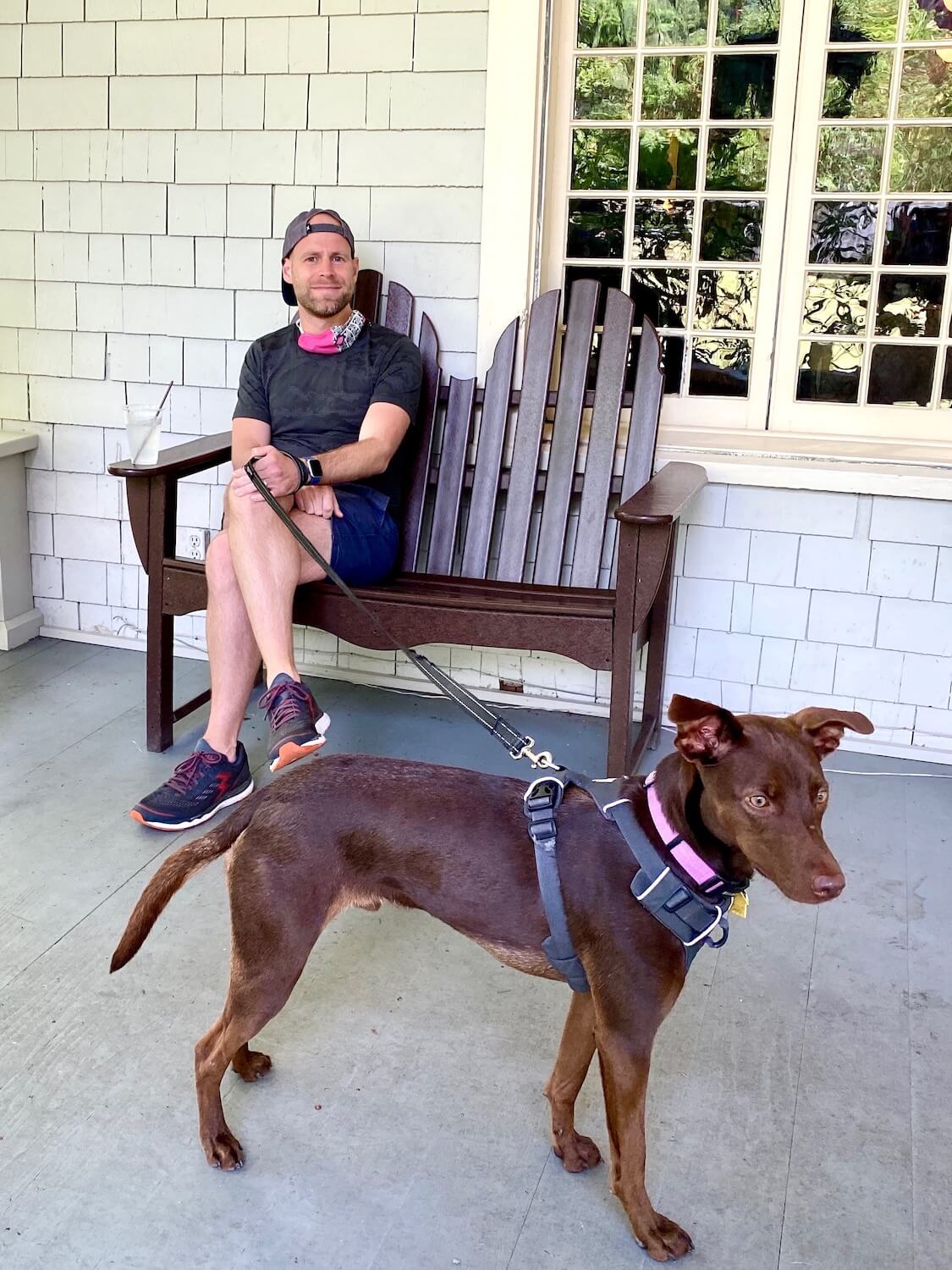 A man sits on a bench on the porch of the Lake Crescent Lodge holding a leash to a sleek brown dog with tall legs and a shiny, healthy looking coat.