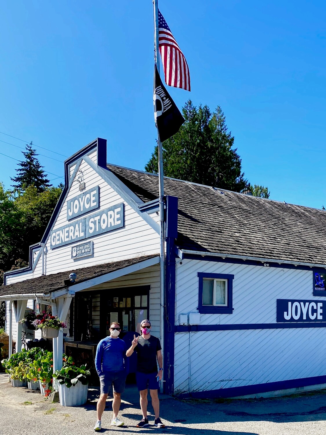 The Joyce General Store is a rustic, timeless small town meeting place. The pioneer style building is gray wood panels with bright blue trim and two men stand out from wearing face masks as a flagpole stands tall with an American flag and a black POW flag. The sky is blue and a green leaved maple tree rises up behind the age old store.