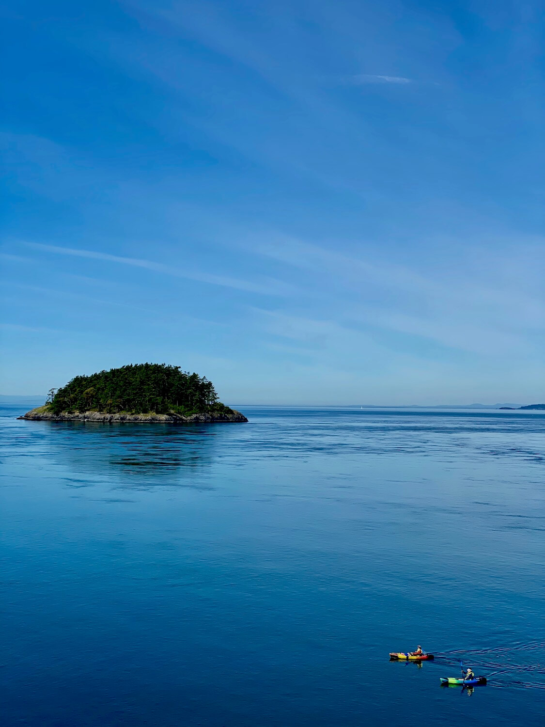 The views from Deception Pass are beautiful on this day trip to nature.  In this shot, Deception Island sits in the middle of the Salish Sea, rocky shore and short fir trees huddled together on the small parcel of land.  In the foreground two kayakers glide by with paddles in hand. 