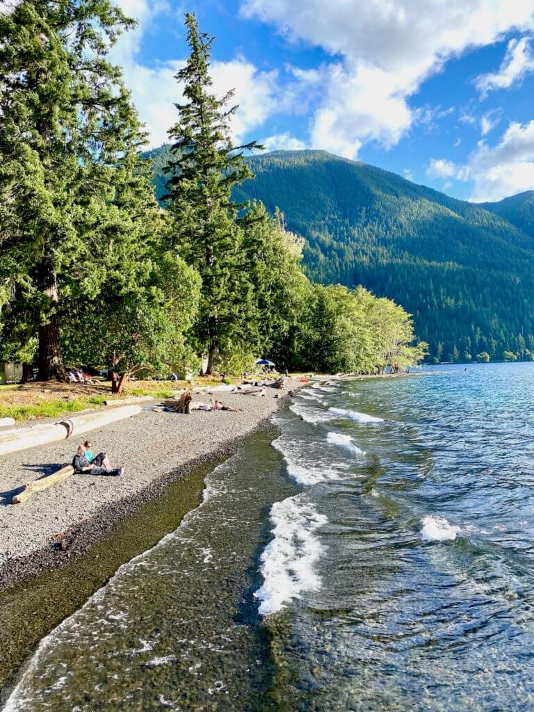 Every day is a pleasant day lake side at the Lake Crescent Lodge. In this shot light lake waves make white crests as they swoosh up on the pebbly beach where a couple leans against a log. Other people are farther down the beach also leaning against logs as fir trees rises up along the bank. In the distance high mountains, tree-lined rises up to the cloud patched blue sky.