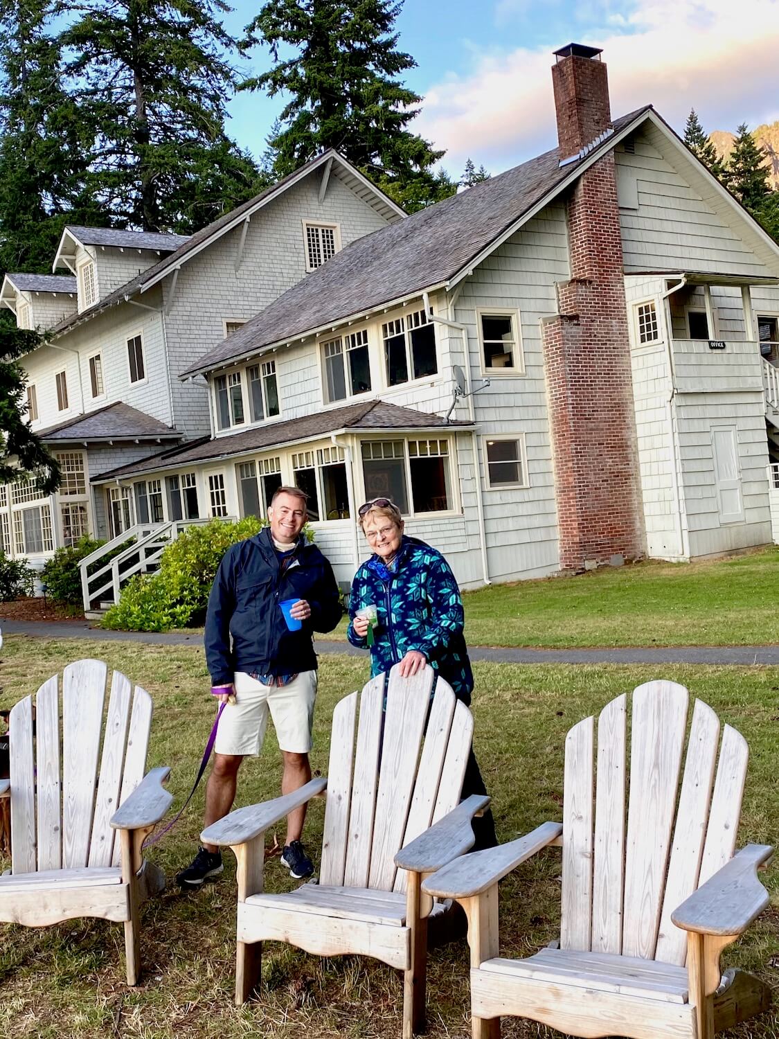 The outside of Lake Crescent Lodge in amongst nature. These two lodge visitors are enjoying a beverage next to three oversized Adirondack chairs. The sky is changing to sunset colors with a brush of pink across the otherwise blue, tree lined sky.