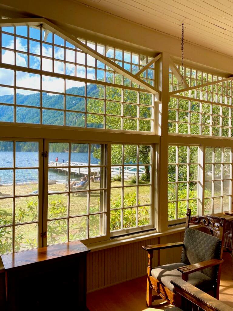 The view looking out of the Lake Crescent Lodge sunporch toward the lake and dock. They windows offer a full view and are made up of tiny panes of glass. Inside the porch is a antique brown chair.