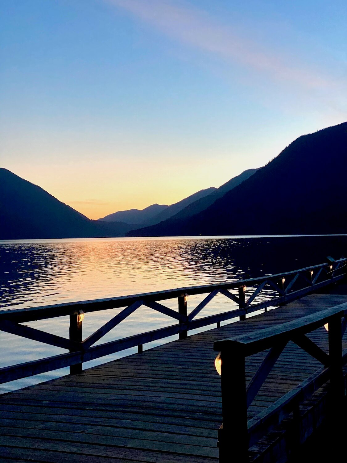 Lake Crescent aglow in sunset hues of peach and powder blue sky reflecting on the shimmering lake. The skyline of the mountains framing the lake are already dark while the dock that protrudes into the glacial water has faint lights affixed to each post holding up the criss crossed railing.
