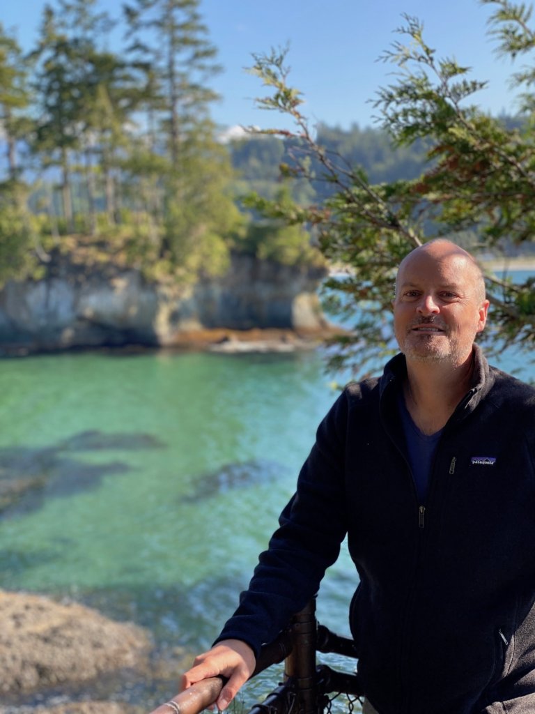 A selfie of Matthew Kessi on the stairs down to a rocky coastline on the Olympic Peninsula. He is wearing a black Patagonia Jacket and the sea water in the distance is a low tide clear green color while the sky is blue.