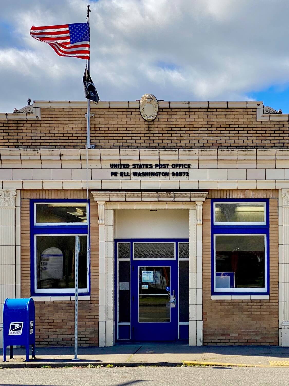 The quaint and tiny Pe Ell, Washington post office is built from light orange bricks with stone archway and blue trim around two windows. A sharply painted blue USPS post office box sits on the sidewalk next to a flag pole flying the US glad and a POW flag.