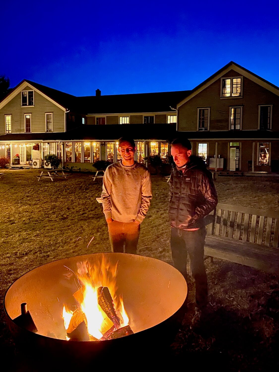 Two men stand beside a fire pit in front of a historic looking wood framed hotel. The fire is bright and crackling yellow and reflects on the faces of the two men. In the background, the hotel commands the skyline, ablaze with lights shining from within all under a blue dusk sky.