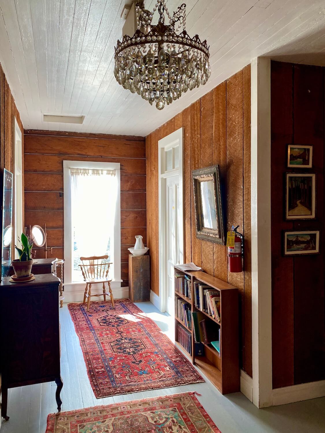 The upstairs hallway of the Tokeland Hotel has walls made from vertical fir boards and white painted ceiling. An ornate crystal chandelier hands in the crossing of two hallways and there are assorted antique furniture pieces. There are two oriental rugs on the floor and bright light emitting from the outside.