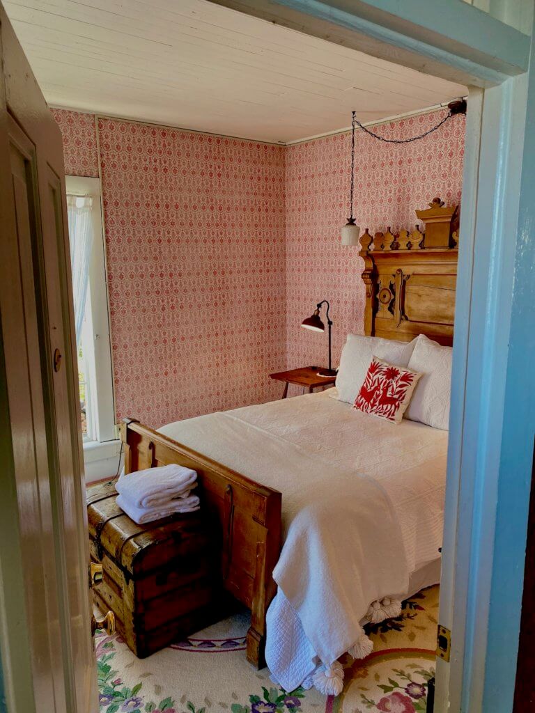 Room #1 at the Tokeland Hotel, on the Washignton Coast. This view is looking into the room from the hallway. The room has wallpaper with bright red geometric circles and a white wood slat roof. The bedspread is white and the ornate headboard made from a walnut looking wood.