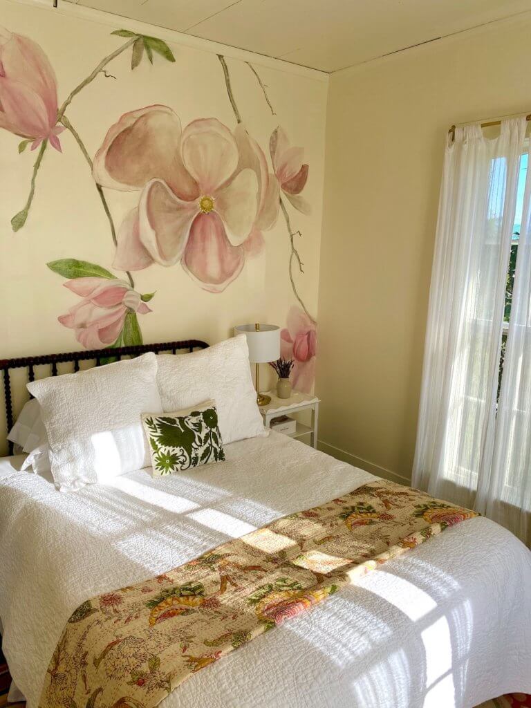 This photo of a room at the Tokeland Hotel on the Washington Coast shows a light filled room with a queen sized bed with cream colored walls and a magnolia tree branch and pink flower etched on the wall in water colors.