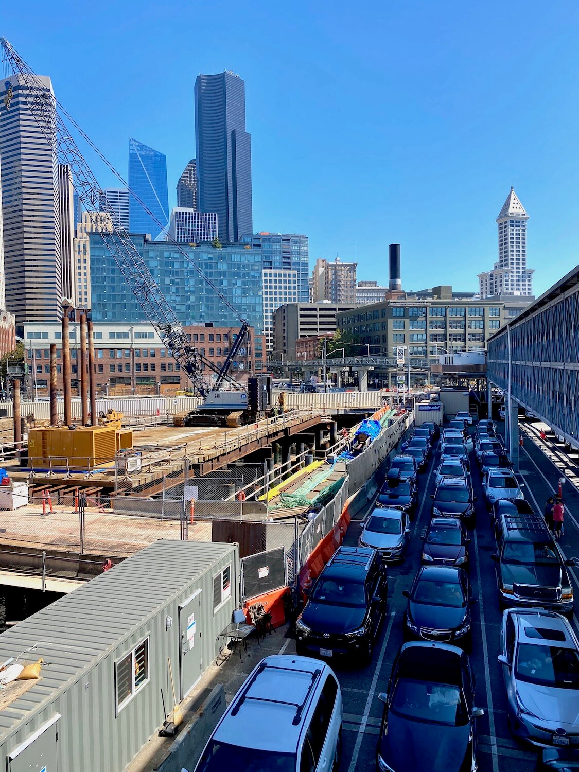 The Coleman Dock in Downtown Seattle has cars lined up to drive onto a Washington State Ferry. There are three lanes of cars and they are sandwiched between heavy construction and the long passenger walkway out to the boats. In the background the Columbia Center rises high into the Seattle skyline with iconic Smith Tower with it's pointed triangle white roof.