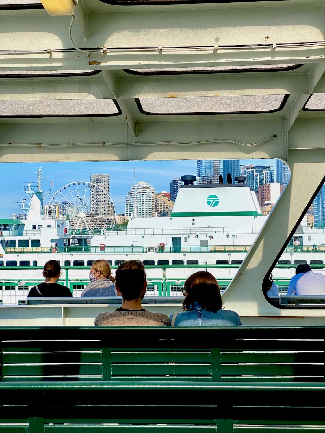 Ferry passengers look out toward Downtown Seattle as the ferry prepares to navigate it's way to a destination on the other side of the Puget Sound. The benches are painted green and another ferry is in the background, just in front of Seattle's Big Wheel attraction downtown.