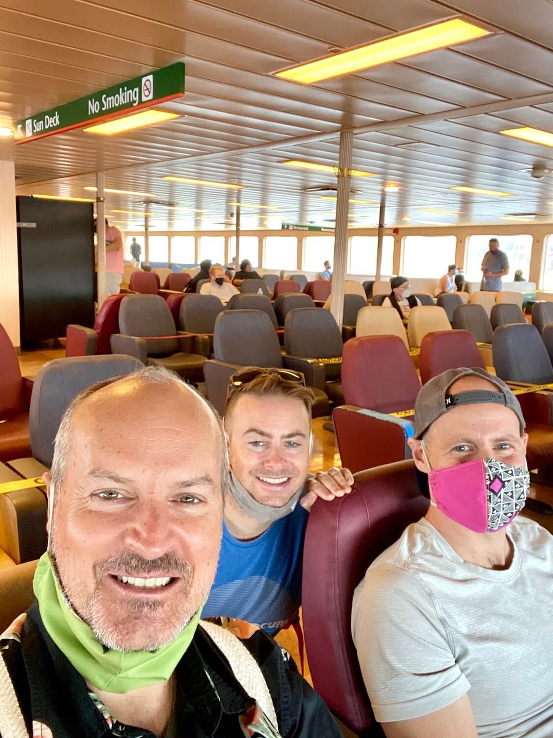 Passengers on the Washington State ferry are navigating their way to a fun destination. There are three men smiling for the camera including Matthew Kessi. The shot is taken indoors on the passenger deck of the ferry and there are hundreds of empty seats behind the men, colored red white and blues.