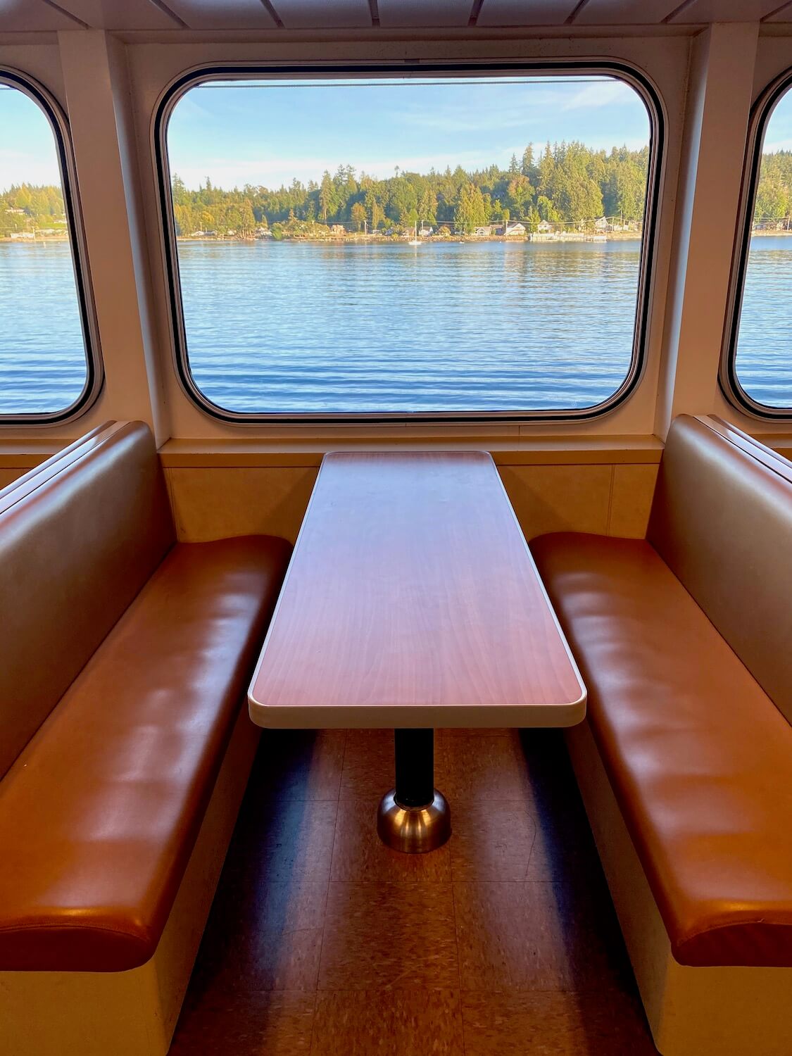 Inside a Washington State Ferry, passenger seating and booths like this one allow views out large picture windows. This shot shows a very clean orange booth with vinyl seating and a faux wood dining table suitable for playing cards or workmen with a computer. The water is gently folding light waves outside and trees and blue sky make up the scenic background.