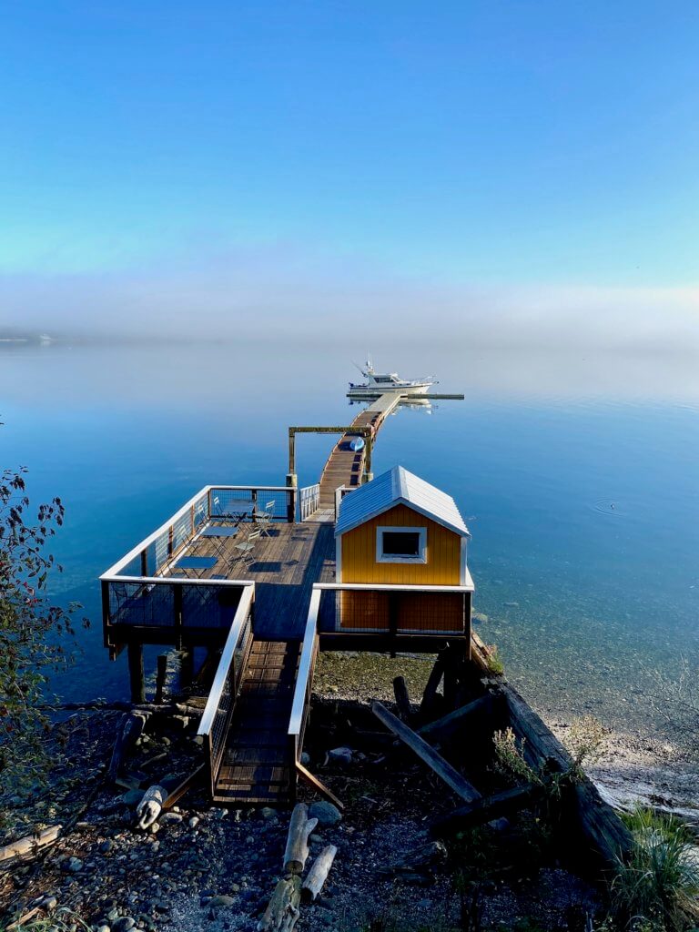 The dock at Captain Whidbey projects into Penn Cover, which in this morning view has fog fading off into the horizon. There is a yellow boat house with white trim on the top larger dock structure and the sky is a bright blue.