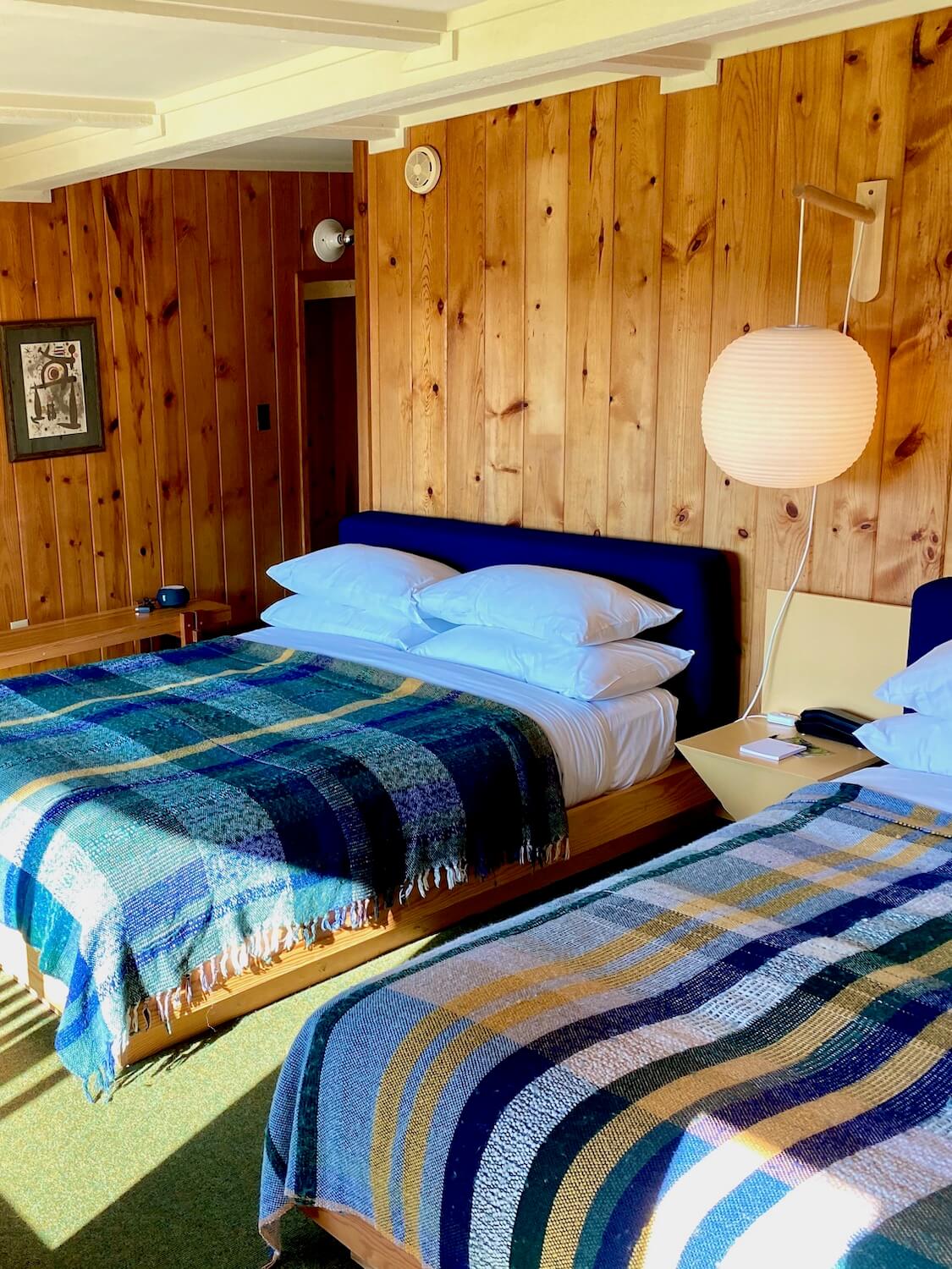Sunlight fills the room of a Lagoon Area building in the Captain Whidbey lodge complex. There are plaid wool blankets draped over the two queen beds with plush white pillows. The walls are a light pine wood and a large Nelson blue like lamp hangs in between the two beds.