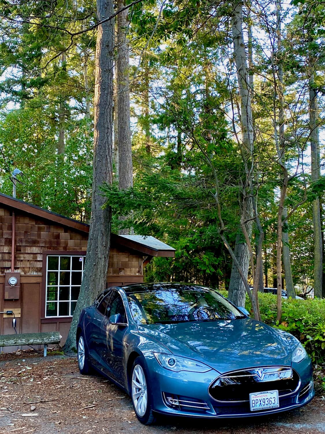 An electric vehicle charges along side a brown building with cedar shake siding. The electric meter is seen next to the box connecting to the vehicle which is situated under coastal fir trees. 