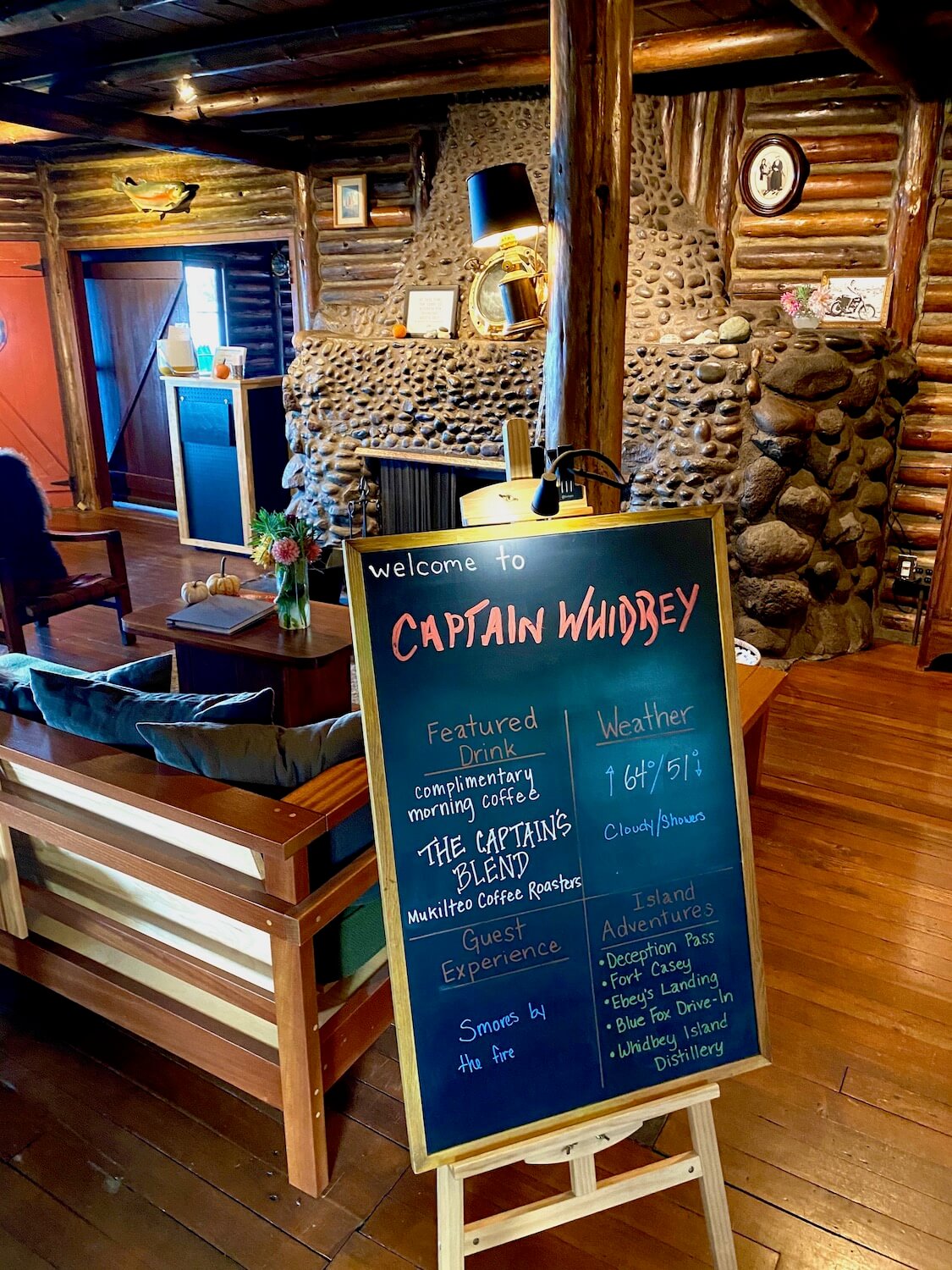 A chalkboard on a wood easel greets visitors to the Captain Whidbey, an inn that sits steps away from Penn Cove on Whidbey Island. The chalk board displays a variety of helpful information. Behind the easel the mixture of wood textures adds a cozy atmosphere against the gray stones that compose the large fireplace.