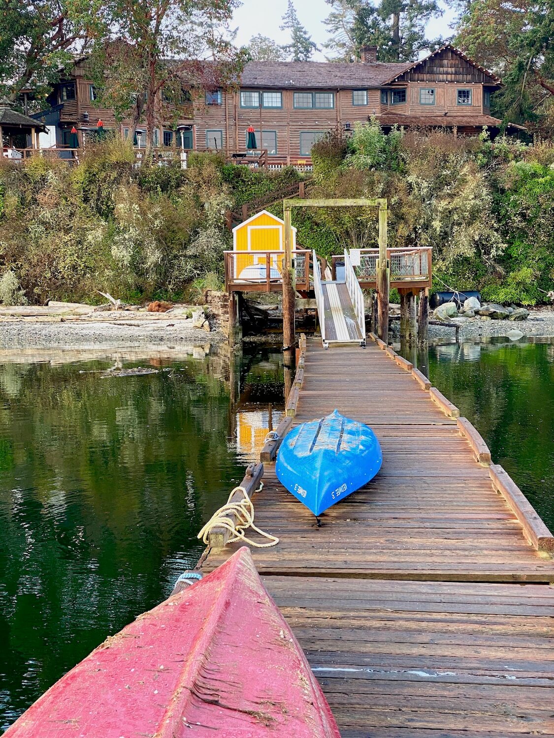Captain Whidbey hotel, from the vantage point of Penn Cove on Whidbey Island. The dock has a red and a blue canoe turned upside down on the dock and in the distance there is a yellow boat house with white trim. The hotel sits above the water in a commanding position.