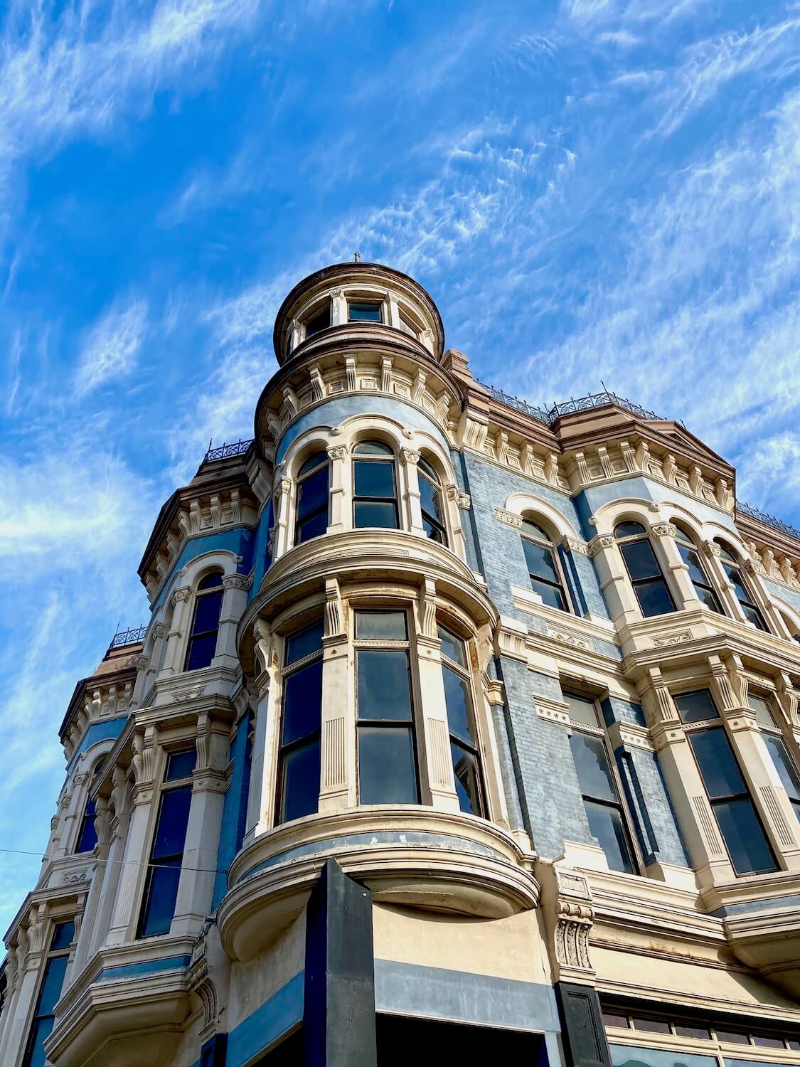 A towering Victorian building rises up on the Main Street of downtown Port Townsend. The ornate three story building utilities a combination of light blue brick with ornate white bordered windows with metal fencing like grating that lines the roof. The sky above is blue with swirls of white clouds.