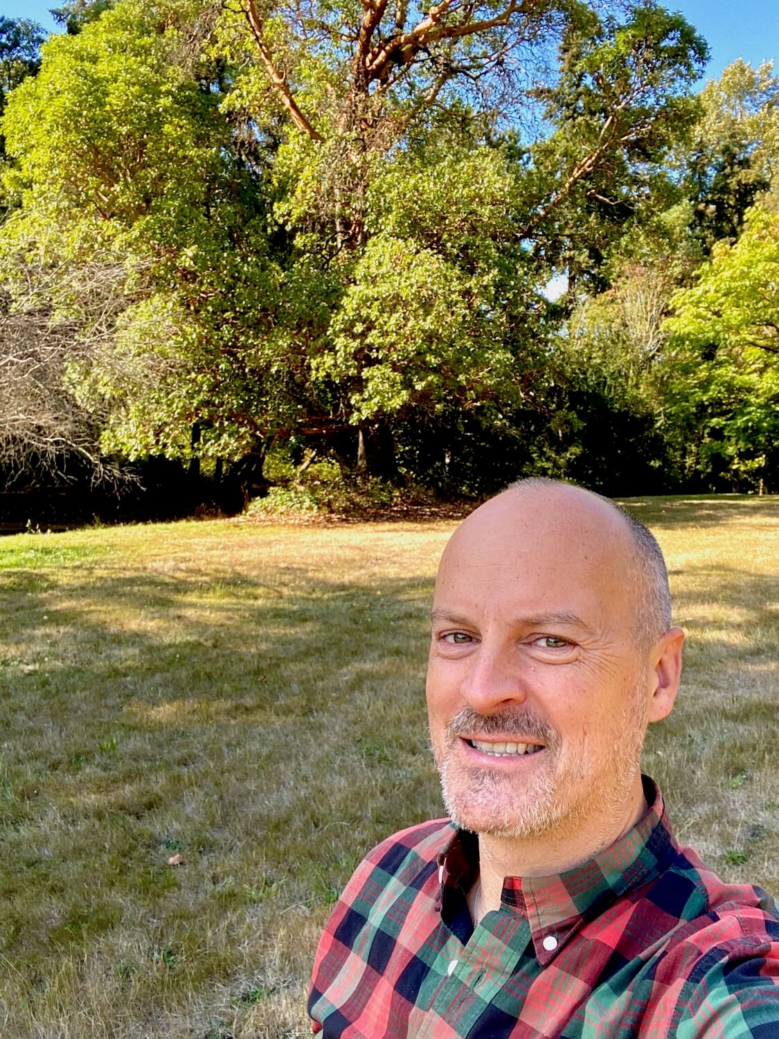 Selfie of Matthew Kessi in an open meadow in a Seattle park.  The trees in the background are full of green leaves against a blue sky and he's smiling and wearing a red, black and green checkered button up shirt.  