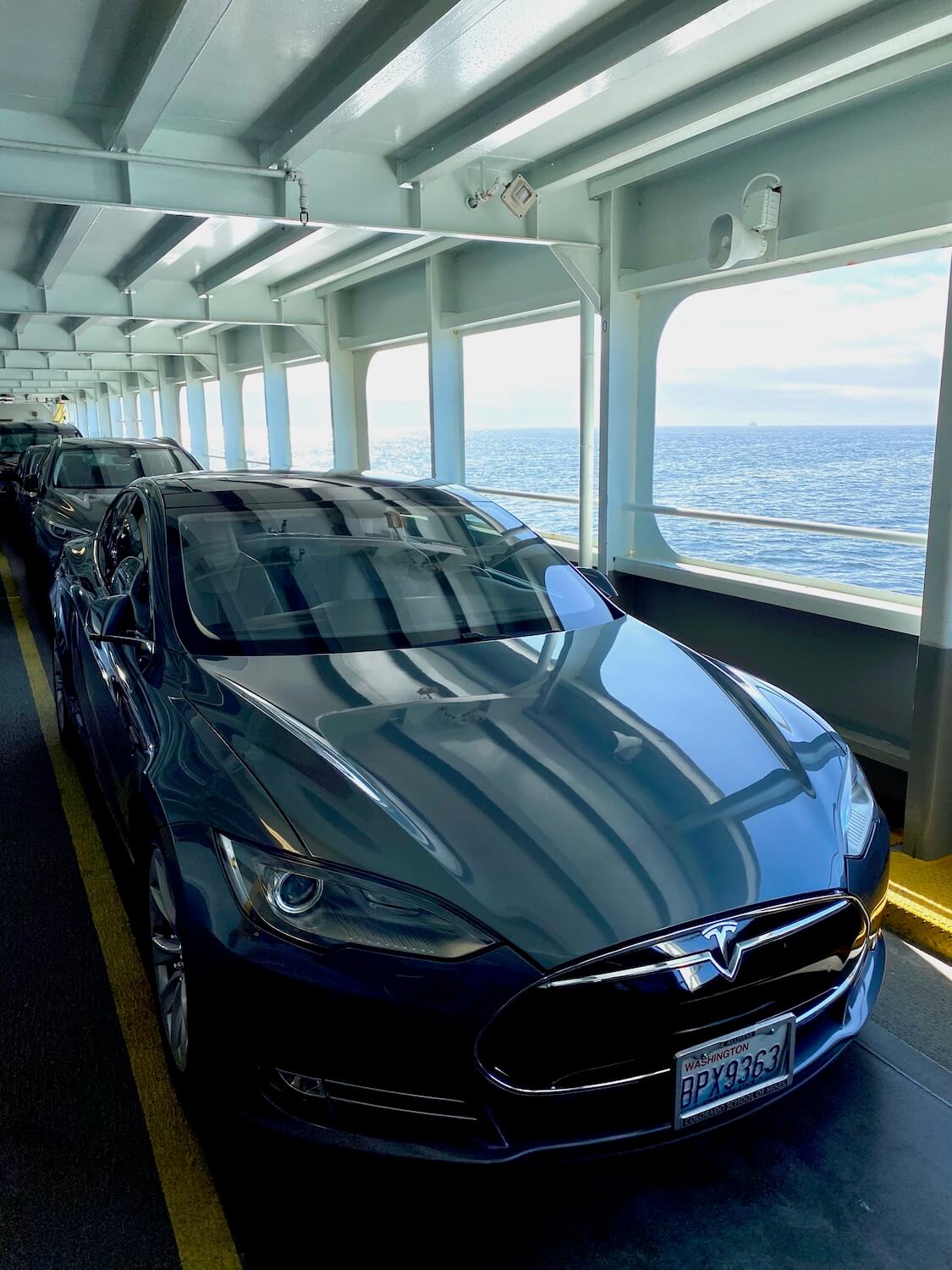 The Tesla beginning a road trip around the Puget Sound, on board a Washington State Ferry.  The car is in line with many others amongst metal beam structures against the flowing water beyond. 