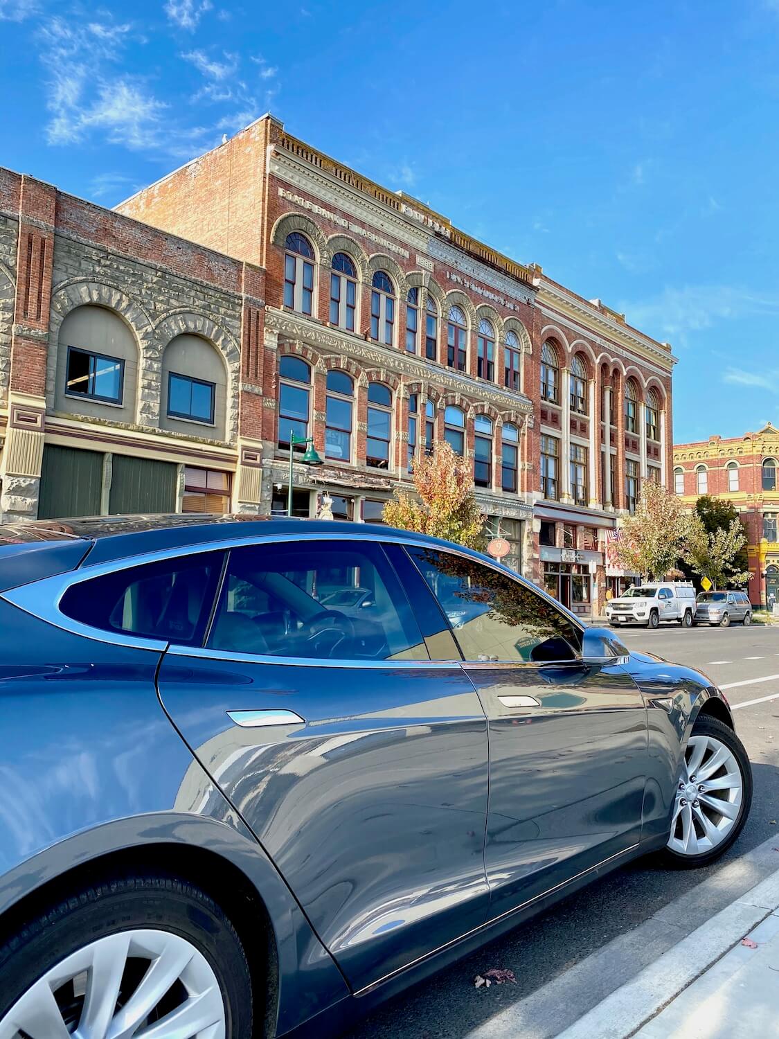 A shiny gray car is parked along the side of the road amongst historic buildings with ornate brick facades.  