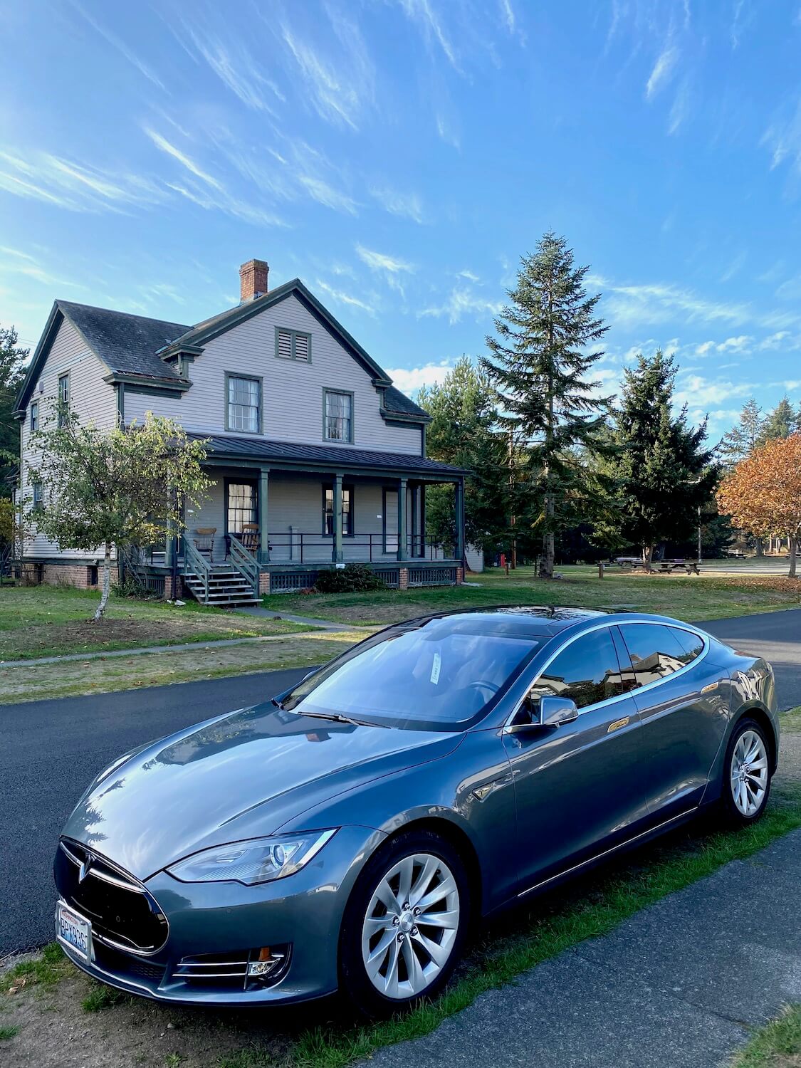 A Tesla is parked on a paved street across from a historic officer home on the grounds of Fort Worden State Park. The house is painted pink with olive green trim and the sky is blue above.  