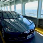 A Tesla Model S sedan sits on the deck of a Washington State Ferry with open Puget Sound waters in the distance against a cloudy sky. The vehicle is a metallic gray blue and shows the prominent Tesla hood ornament on the front. There are other cars behind the Tesla and the metal structure of the ferry are painted white on the ceiling above.