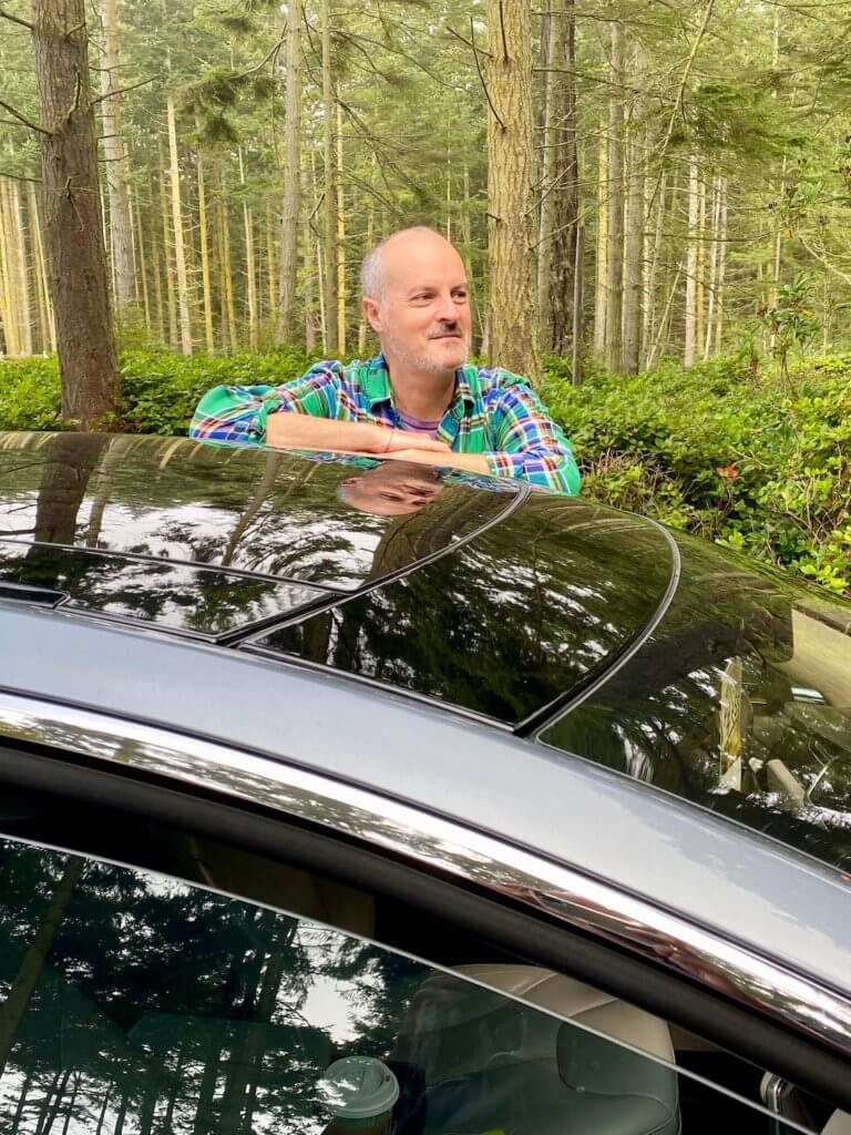Selfie of Matthew Kessi standing outside a Tesla S series in the middle of a woods scene. The trees in the background are fir with variegated bark and he is wearing a green plaid shirt while resting his arms on the shining metallic blue sedan.