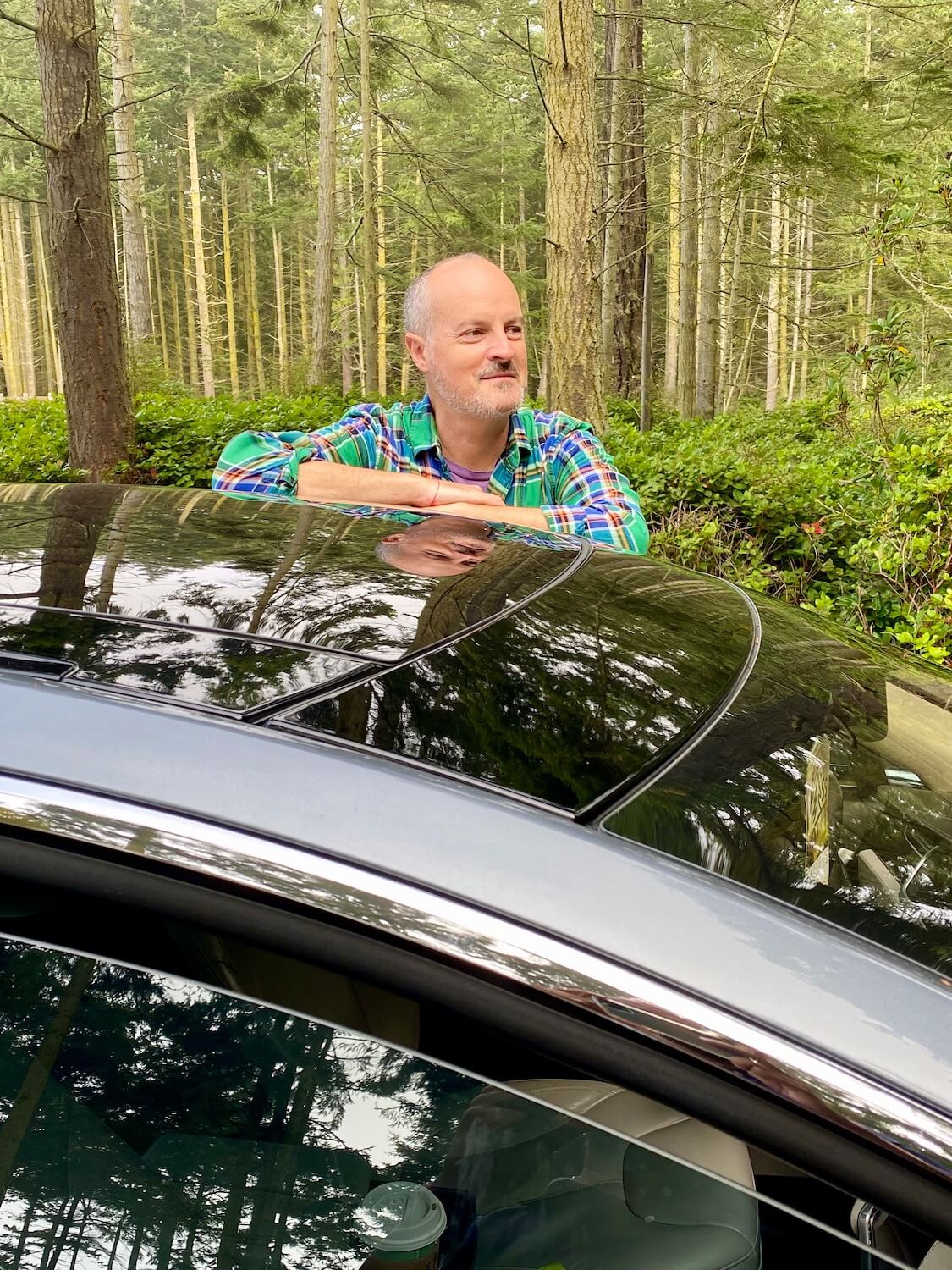Selfie of Matthew Kessi standing outside a Tesla S series in the middle of a woods scene. The trees in the background are fir with variegated bark and he is wearing a green plaid shirt while resting his arms on the shining metallic blue sedan.