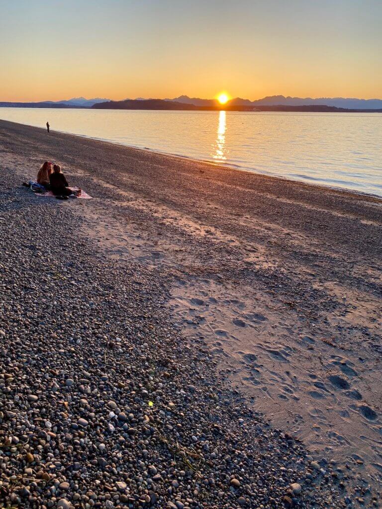 An orange glowing sun sets behind the Olympic Mountains as the rays shine on the vast Puget Sound waters. A couple enjoys the sunset on a blanket on the beach. This is a great winter outdoor thing to do in Seattle.