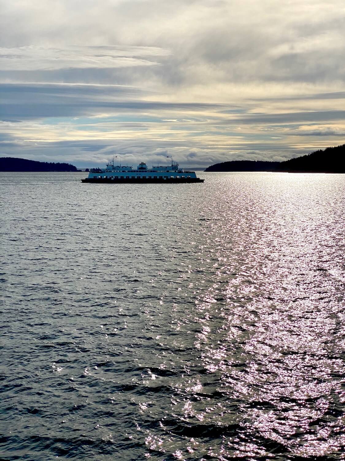 A Washington State Ferry crosses the Puget Sound in between several islands.  The two level ferry is white with a darker color on the base near the water and the see has small choppy waves that glow with the winter sky hosting a sunset. 