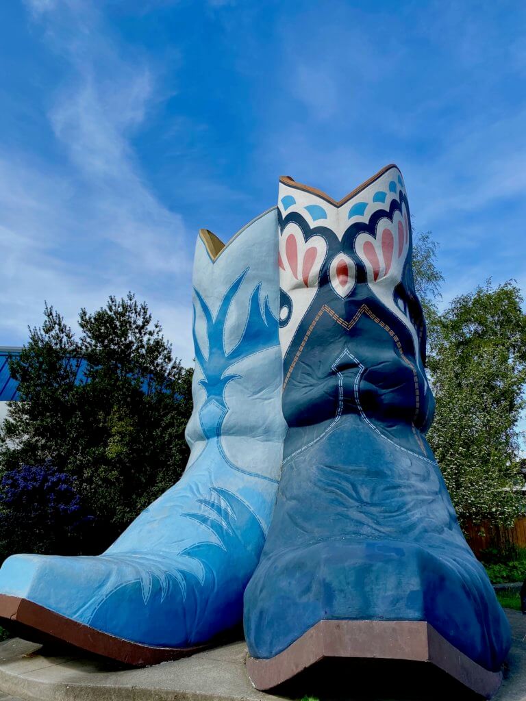 Two large cowboy boots are displayed in a park.  They are various shades of blue and showcase intricate painted stitching in the folds and textures of concrete.  