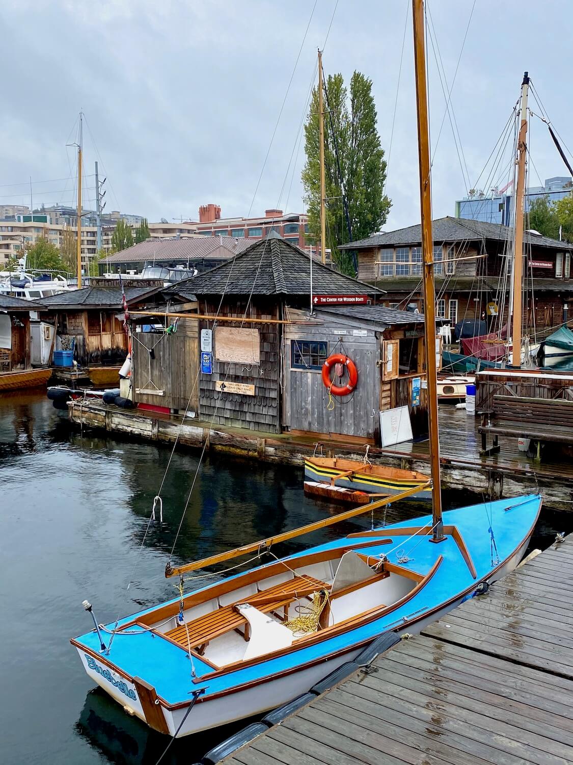 A small blue sailboat is tied to a floating dock, wet and glistening from a recent rain.  In the background are other floating buildings with wood shakes and other marine gear under a blue sky. 