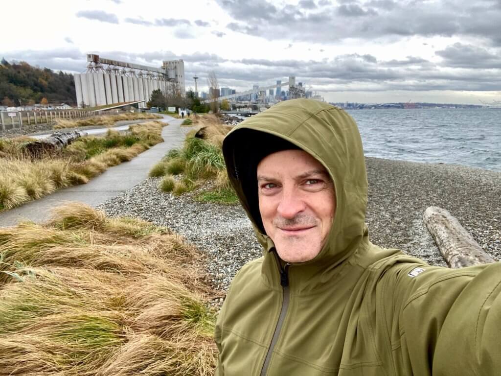 Matthew Kessi stands along the banks of the Salish Sea in Seattle, Washington wearing a green rain jacket with the city in the background.