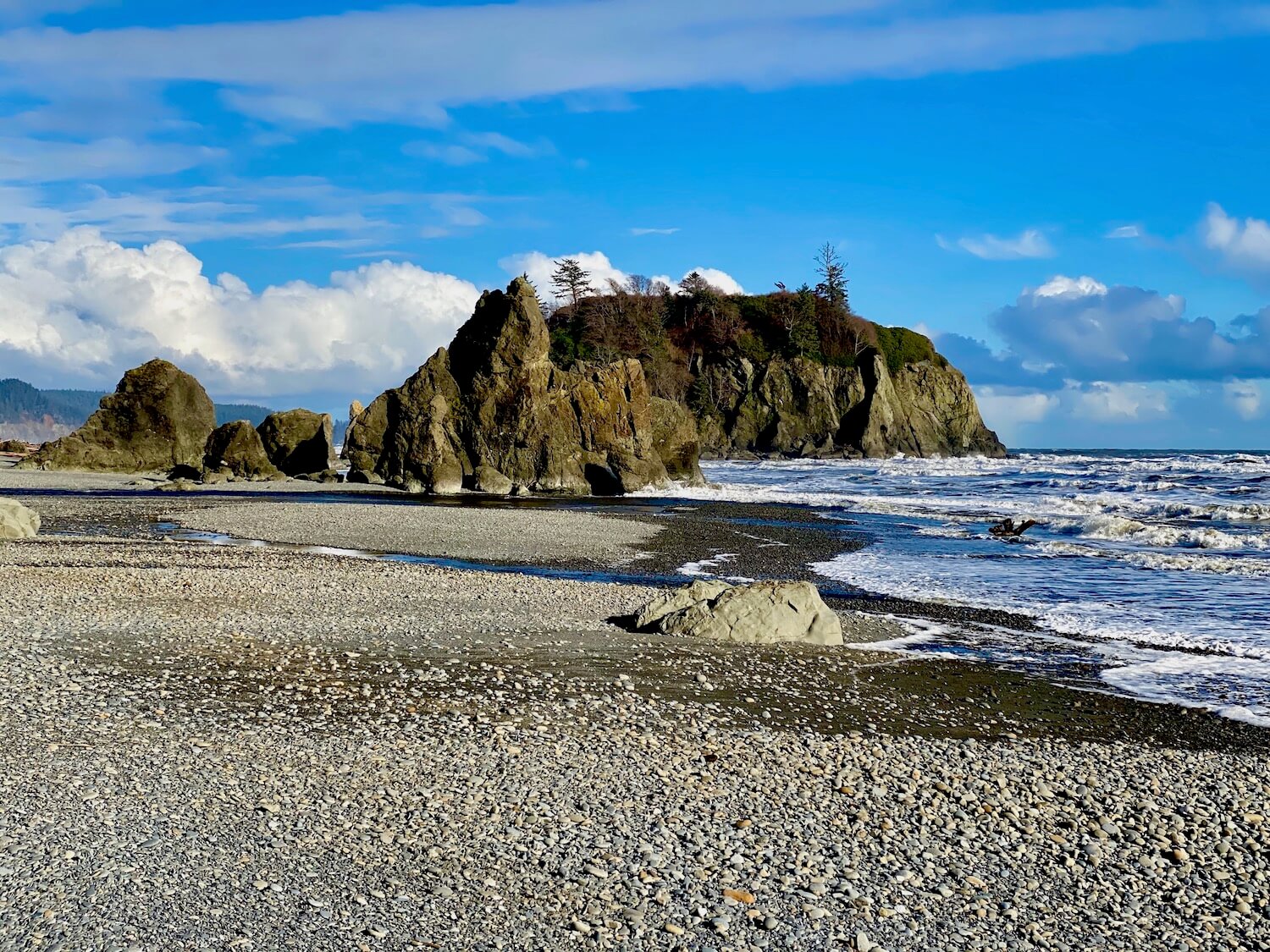 A beautiful beach scene combines bright blue skies with some puffy white clouds and rock stacks indicative of the Washington Coast. The surf is crashing waves onto the rocky beach revealed by low tide.