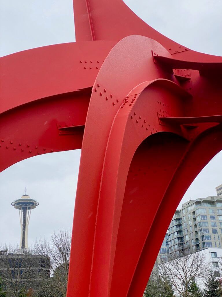 A bright red sculpture bends in a way that allows the iconic Seattle Space Needle to rise up in the background. The red structure is put together with a series of rivets holding the steel together.